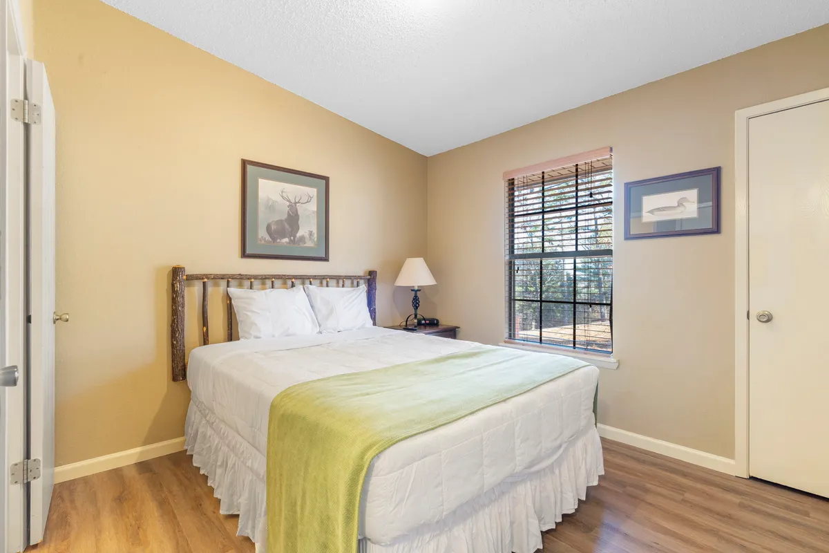 A tranquil queen bedroom with crisp white linens, a green throw blanket, a bedside lamp, window with blinds, and framed artwork. The space radiates comfort and simplicity, ideal for restful nights after outdoor adventures.
