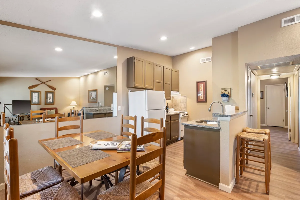 A warm scene showing the dining table adjacent to the kitchen area with open access to the living room. The setting suggests togetherness, shared meals, and easy flow between spaces, with natural light enhancing the inviting mood.