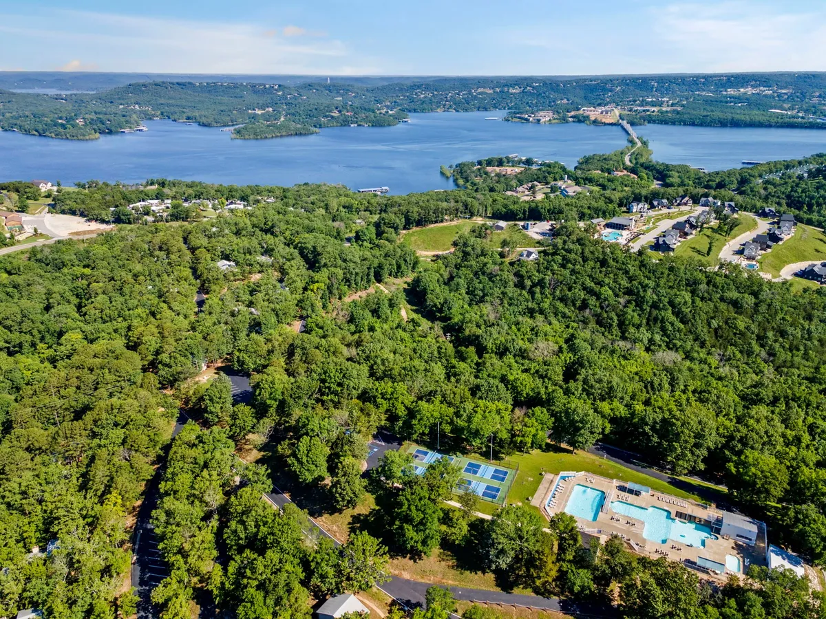 A vibrant exterior shot featuring multiple swimming pools, tennis courts, a sparkling lake, and lush surrounding greenery. The image captures various areas for recreation, including the pool and courts, with the lake as a scenic backdrop. People are visible enjoying the amenities, suggesting active use.