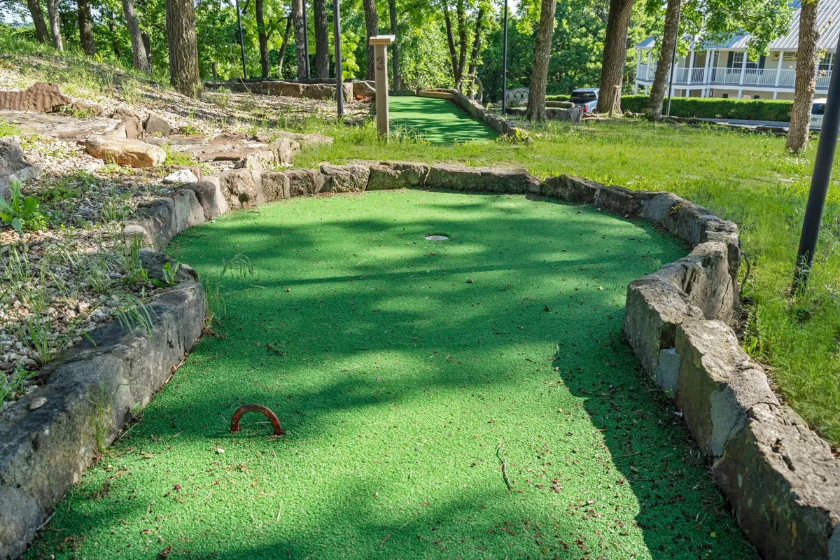 A cheerful mini-golf course is nestled among mature trees, bordered by stone edges. The scene depicts an inviting family-friendly atmosphere, ideal for making memories and enjoying outdoor games.