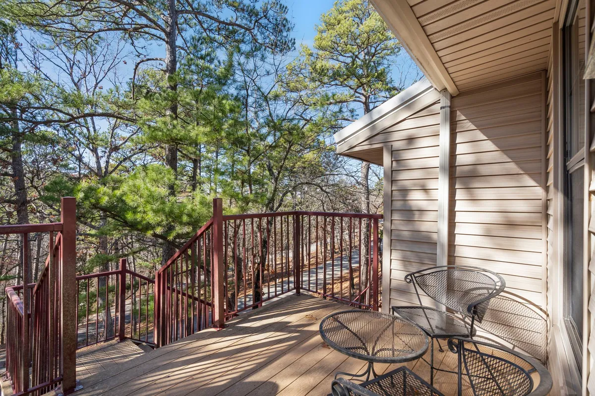 An exterior view of a wooden deck with a metal railing, surrounded by lush trees and outdoor chairs. The setting is tranquil, ideal for enjoying sunrise coffee, sunset toasts, or simply soaking in nature.