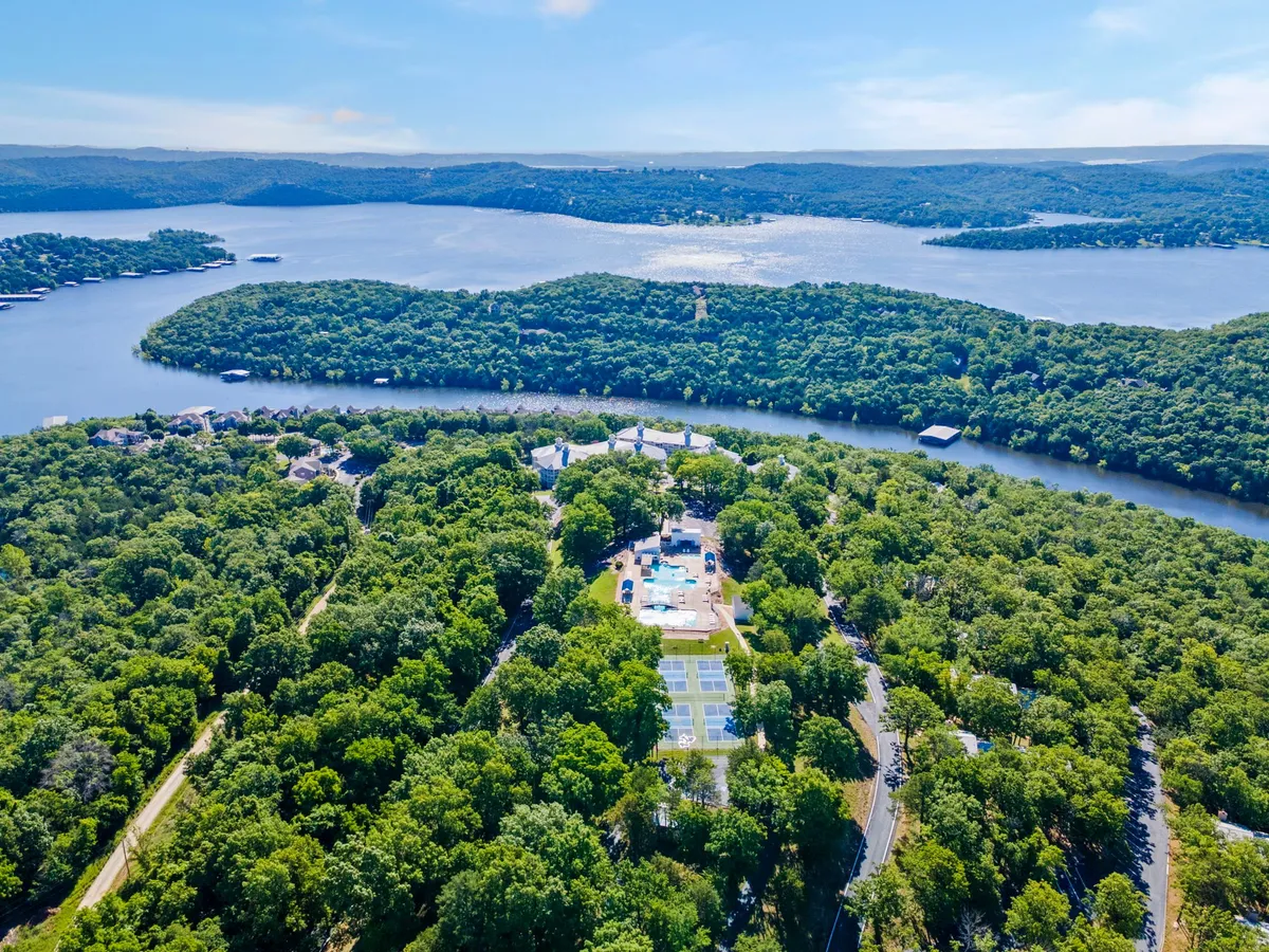 This image showcases a vibrant outdoor recreation area with a sparkling swimming pool, adjacent tennis court, lush green trees, and a beautiful waterfront view in the background. The space appears lively and inviting, perfect for both relaxing and engaging in outdoor activities.