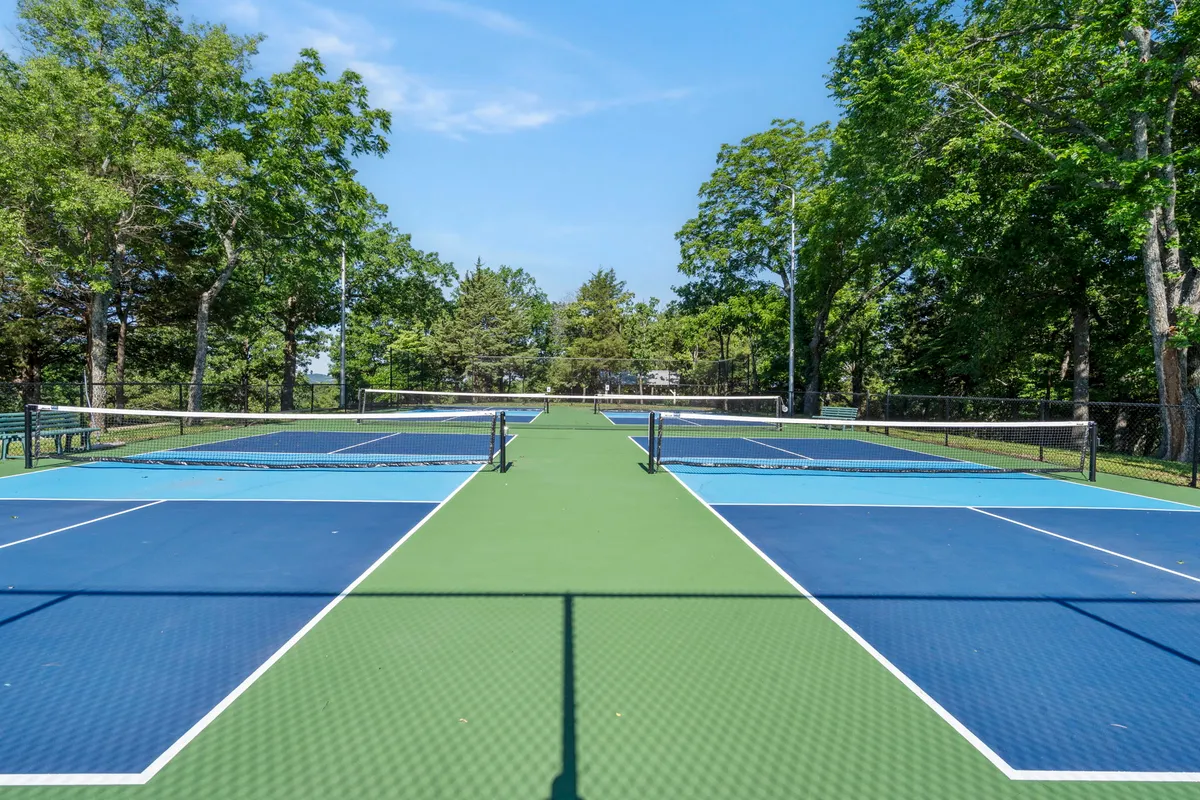 This image shows tennis courts with netting, bordered by tall trees, and the lively environment of people playing and socializing. The bright, energetic scene underlines the active recreation available at Evergreen Ridge.